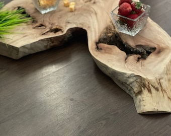 Puede incluir: Una bandeja de servir de madera con un acabado de grano de madera natural. La bandeja tiene una forma única con una gran sección central irregular. Dos pequeños cuencos de vidrio se colocan en la bandeja, uno que contiene queso en cubos y el otro que contiene fresas frescas.