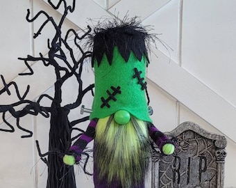 Peut inclure: Un gnome d'Halloween vert et violet avec une barbe noire et verte, des points de suture sur le visage et une araignée sur le corps. Le gnome est debout à côté d'une pierre tombale sur laquelle est inscrit "RIP".