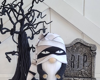 May include: A black and white Halloween gnome wearing a mummy costume with a black eye mask. The gnome is standing in front of a grey tombstone with the word "RIP" on it.