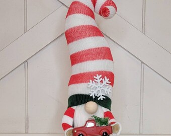 Peut inclure: Un gnome à rayures rouges et blanches avec un chapeau vert et un flocon de neige blanc. Le gnome tient un camion jouet rouge avec un petit arbre vert à l'arrière.