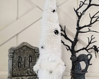 May include: A Halloween gnome figure with a white, wrapped body and a long white beard. Black spiders are attached to the gnome. A tombstone with "RIP" and a black cat statue are also in the image.