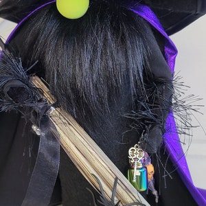 Peut inclure: Un gnome de sorci&egrave;re noir avec un chapeau violet et noir, un nez vert et un balai. Le gnome porte une cape violette et a une petite bouteille avec un charme de cl&eacute; qui pend.