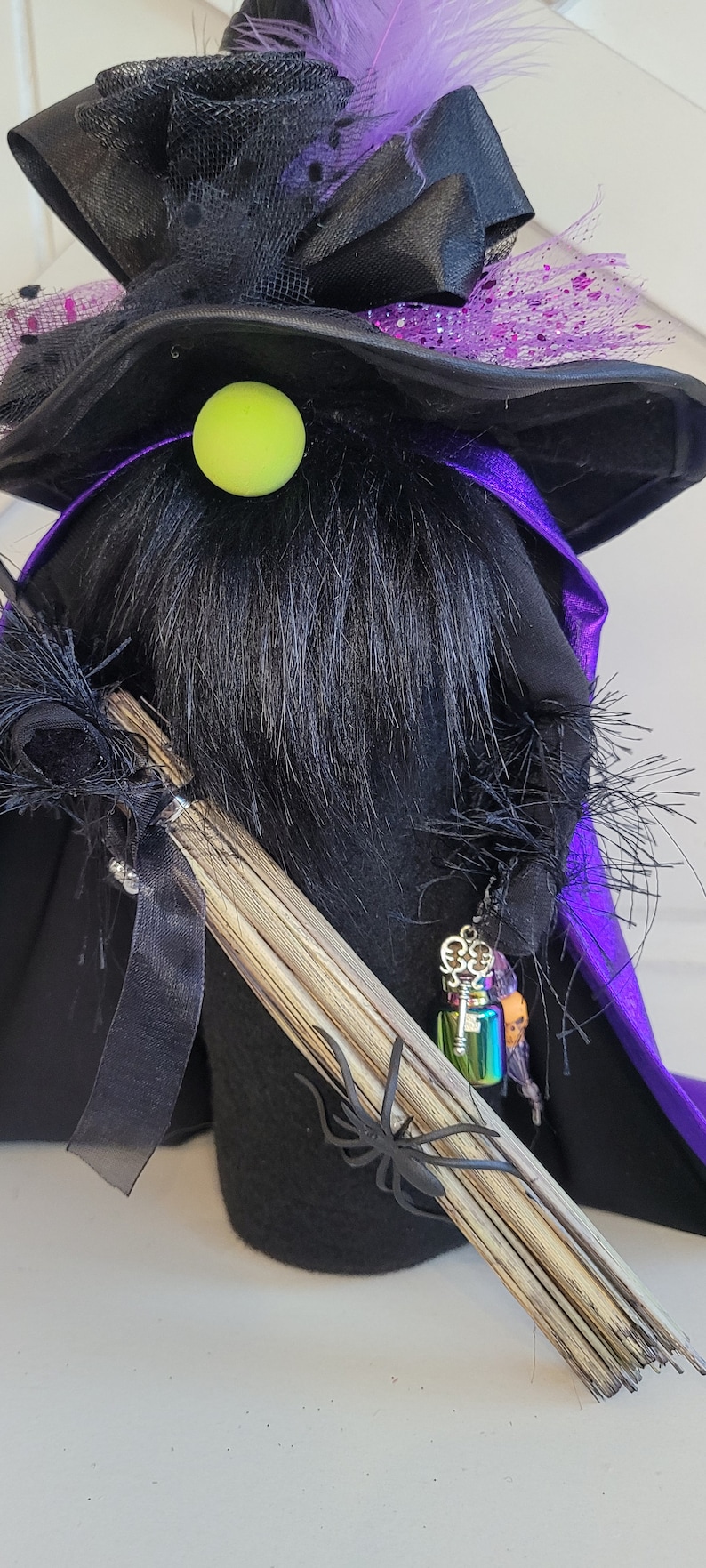 Peut inclure: Un gnome de sorci&egrave;re noir avec un chapeau violet et noir, un nez vert et un balai. Le gnome porte une cape violette et a une petite bouteille avec un charme de cl&eacute; qui pend.