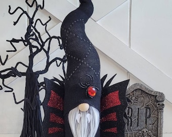Peut inclure: Un gnome d'Halloween noir et rouge avec un long chapeau noir, une barbe blanche et des ailes en paillettes rouges. Le gnome a un œil en gemme rouge et des crocs blancs. Le gnome est debout devant une pierre tombale grise avec le mot "RIP" dessus.