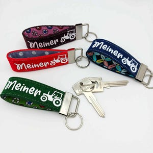 Könnte beinhalten: Vier Schlüsselanhänger aus Stoff in Rot, Grün, Marineblau und Braun, jeder mit einem anderen Muster und dem Wort "Meiner" in Weiß. Eine weiße Traktorgrafik ist ebenfalls vorhanden. Zwei Schlüssel liegen neben dem grünen Schlüsselanhänger.