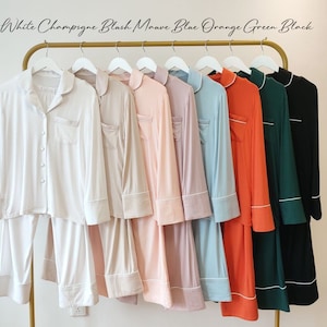 May include: Nine long-sleeved pajamas in various colors, including white, champagne, blush, mauve, blue, orange, green, and black. The pajamas have a collared neckline and a button-up front. Each pajama set has a white trim around the collar, cuffs, and pockets.