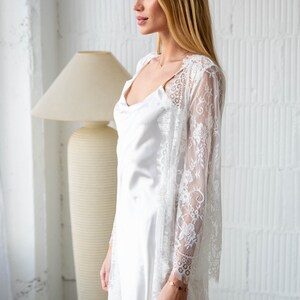 May include: A white lace robe with a satin slip underneath. The robe has long sleeves and a flowing silhouette. The model is wearing a gold bracelet on her wrist.