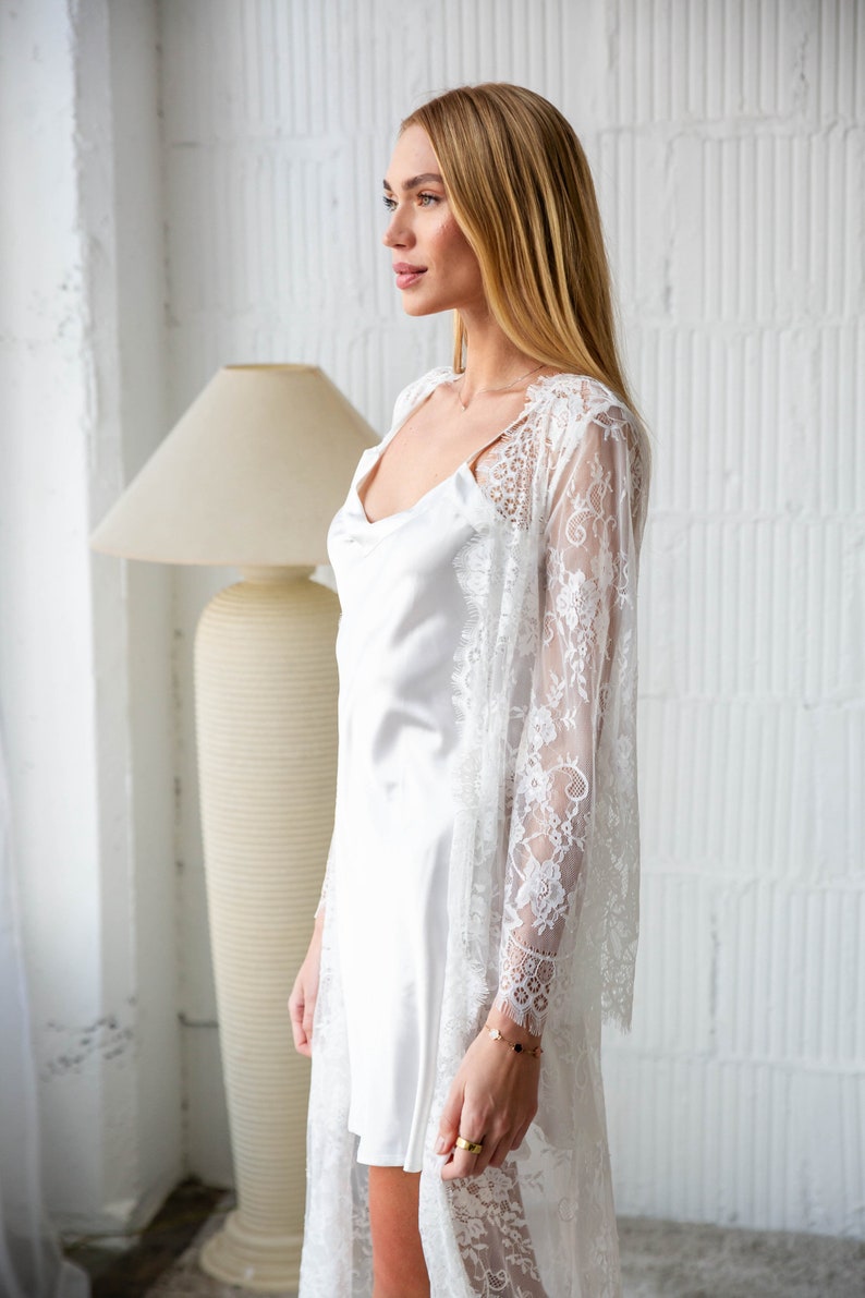 May include: A white lace robe with a satin slip underneath. The robe has long sleeves and a flowing silhouette. The model is wearing a gold bracelet on her wrist.