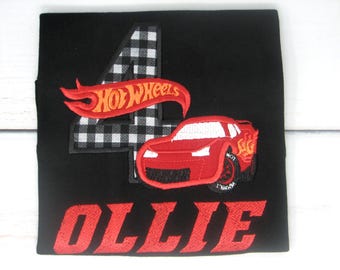 May include: Black t-shirt with a red and white checkered number four, a red race car with flames, and the words "Hot Wheels" in orange. The name "Ollie" is embroidered in red below the car.