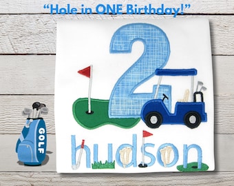 May include: White t-shirt featuring a blue golf cart, a green putting green, and the name "Hudson" in blue. The number "2" is also in blue. A golf bag with the word "GOLF" is on the left. The text "Hole in ONE Birthday!" is at the top.