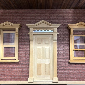Peut inclure: Trois cadres de fenêtres et de portes en bois avec des moulures décoratives, placés contre un mur de briques rouges. Les cadres sont peints d'une couleur bois clair et ont un design simple. Les fenêtres ont des vitres claires et la porte a un design à six panneaux.