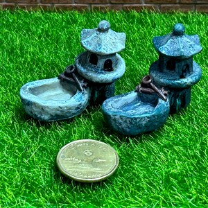 May include: Two miniature gray stone pagodas with a small, gray stone basin in front of each. The pagodas have a tiered roof and are decorated with small windows. The basins are shallow and have a rounded shape. The pagodas and basins are sitting on a green artificial grass surface.