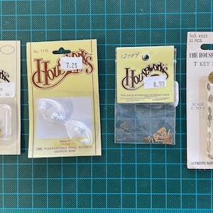 May include: Three packs of miniature brass hardware for dollhouses. The packs are labelled "Houseworks" and include a range of pieces, such as hinges, door knobs, and T-key hole plates. The packs are on a green grid background.
