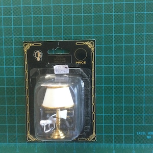 May include: A miniature gold table lamp with a white shade and a cord. The lamp is packaged in a clear plastic blister pack with a black and gold cardboard backing. The packaging includes the text "Excel Hobby Blades No. 50002".