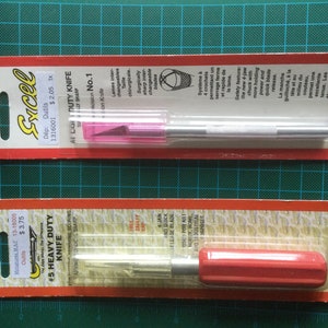 Peut inclure: Deux couteaux de bricolage emballés. L'emballage supérieur est étiqueté "Excel AI Light Duty Knife No. 1" avec un protège-lame rose. L'emballage inférieur est étiqueté "#5 Heavy Duty Knife" avec un manche rouge.