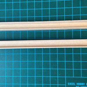 May include: Two wooden strips with a rectangular cross-section. The strips are light brown and have a smooth surface. The strips are lying on a green cutting mat.