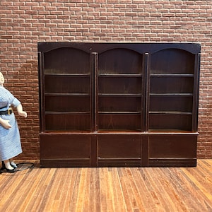Peut inclure: Une bibliothèque miniature en bois brun foncé avec trois sections et plusieurs étagères. Une poupée en robe bleue et blanche se tient à gauche de la bibliothèque. L'arrière-plan est un mur de briques.