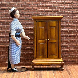 Peut inclure: Une armoire en bois miniature à deux portes et une figurine d'infirmière vêtue d'une robe à carreaux bleus et blancs. La figurine porte une coiffe blanche et des chaussures noires. Le fond est un mur de briques.