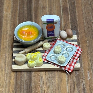 May include: A miniature baking set with a wooden chopping board, a bowl of orange liquid, a rolling pin, a bag of Gold Medal All-Purpose Flour, a muffin tin with dough, and two eggs.