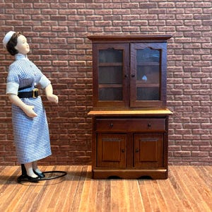Peut inclure: Un meuble miniature en bois avec des portes vitrées et des tiroirs, à côté d'une poupée vêtue d'une robe à carreaux bleus et blancs et d'un bonnet blanc. Le meuble est brun foncé, et la poupée est sur une surface en bois.