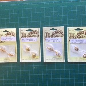 May include: Four packs of gold-coloured decorative embellishments, each with a different design. The packages are labelled "Houseworks LTD" and include the text "Dép: Quincaillerie". The embellishments are on a green grid surface.