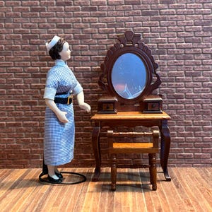 Peut inclure: Une poupée miniature vêtue d'une robe à carreaux bleus et blancs et d'une coiffe blanche se tient à côté d'une coiffeuse en bois avec un miroir ovale. La coiffeuse a deux petits tiroirs et un tabouret assorti. Le fond est un mur de briques.