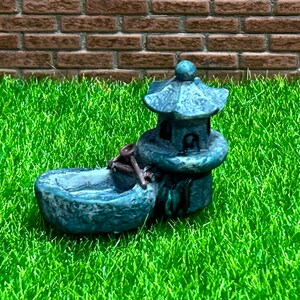 May include: A miniature gray stone pagoda with a small boat-shaped basin in front. The pagoda has a decorative design and the basin has a small wooden paddle.