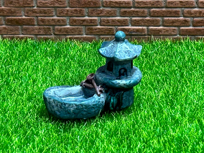 May include: A miniature gray stone pagoda with a small boat-shaped basin in front. The pagoda has a decorative design and the basin has a small wooden paddle.