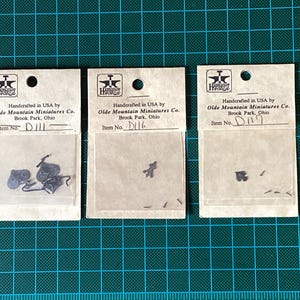 Peut inclure: Trois petits sacs en papier brun avec du texte noir. Chaque sac contient une petite pièce en métal. Le texte sur les sacs indique "Handcrafted in USA by Olde Mountain Miniatures Co. Brook Park, Ohio". Les numéros d'articles sont D111, D116 et D117.