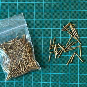 May include: A clear plastic bag filled with a large quantity of small, gold-coloured tacks. A smaller pile of tacks is scattered on a green cutting mat.