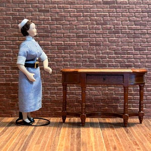 Peut inclure: Une poupée miniature vêtue d'un uniforme d'infirmière bleu et blanc à carreaux, d'une coiffe blanche et de chaussures noires, se tient à côté d'une petite table en bois avec un tiroir. Le fond présente un mur de briques et un sol en bois.
