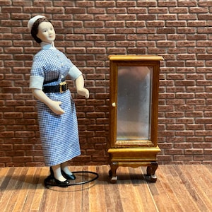 Peut inclure: Une poupée miniature vêtue d'un uniforme d'infirmière à carreaux bleus et blancs, coiffée d'un bonnet blanc et chaussée de chaussures noires, se tient à côté d'une petite armoire en bois avec une porte vitrée. L'armoire a des détails ornés et est placée contre un mur de briques.