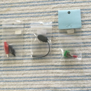 May include: Three small clear plastic bags containing electronic components. The first bag contains two alligator clips, one red and one black. The second bag contains a small black microphone with a wire. The third bag contains a small green toggle switch.