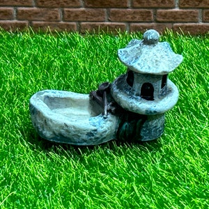May include: A small, gray stone fountain with a pagoda-style roof. The fountain is designed for a miniature garden or fairy garden.