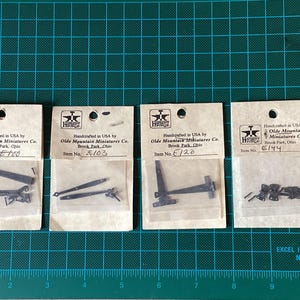 May include: Four small packages containing metal hardware for model railway. Each package has a white label with black text that reads "Handcrafted in USA by Olde Mountain Miniatures Co. Brook Park, Ohio" and an item number. The item numbers are E100, E103, E120, and E144.