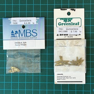 May include: Two packs of brass hinges for model building. The pack on the left is labelled "HINGE-8 2/pk. Fancy Hinges" and is from the company "MBS Model Builders Supply". The pack on the right is labelled "Greenleaf 4 brass hinges (1" Scale)" and is from the company "Greenleaf Products, Inc."