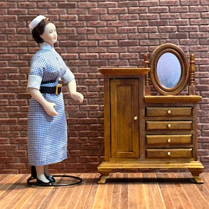 Peut inclure: Une poupée miniature vêtue d'une robe à carreaux bleus et blancs et d'un bonnet blanc se tient à côté d'une coiffeuse en bois avec un miroir. La coiffeuse a des tiroirs et une petite armoire. Le fond est un mur de briques.