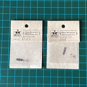 Peut inclure: Deux petits sacs en papier blanc avec du texte noir. Les sacs contiennent de petites pièces métalliques. Le texte sur les sacs indique "Handcrafted in USA by Olde Mountain Miniatures Co. Brook Park, Ohio". Les numéros d'article sont "D115" et "D118".