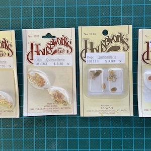May include: Four packs of miniature decorative items labelled "Houseworks LTD" with the text "Dép: Quincaillerie". The packs contain small gold-coloured metal charms and embellishments. The packs are on a green cutting mat.