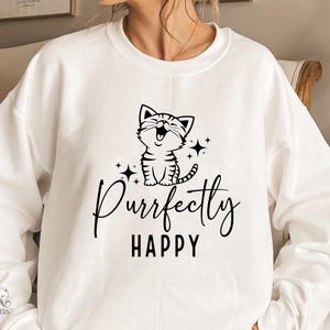 以下が含まれることがあります： 白いスウェットシャツ。黒いグラフィックで、笑顔の猫と「Purrfectly HAPPY」の文字が描かれています。デザインには星が含まれています。カジュアルで快適なスウェットシャツで、気まぐれな猫のデザインです。