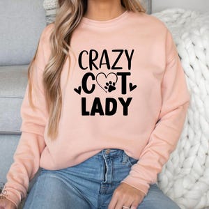 以下が含まれることがあります： 「CRAZY CAT LADY」の黒い文字とハートと足跡のグラフィックが入った、薄いピンク色のスウェットシャツ。スウェットシャツは無地で長袖です。その人は青いジーンズを履いています。