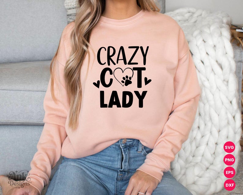 以下が含まれることがあります： 「CRAZY CAT LADY」の黒い文字とハートと足跡のグラフィックが入った、薄いピンク色のスウェットシャツ。スウェットシャツは無地で長袖です。その人は青いジーンズを履いています。