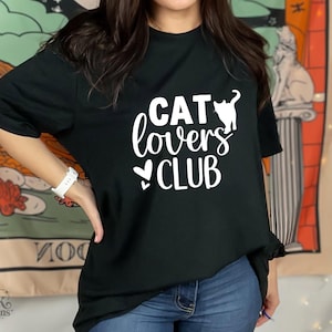 以下が含まれることがあります： 白い「CAT lovers CLUB」の文字と猫のシルエットが入った黒いTシャツ。猫好きにぴったりのカジュアルウェアです。