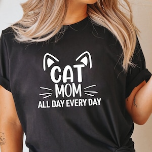 以下が含まれることがあります： 「CAT MOM ALL DAY EVERY DAY」の白文字グラフィックが入った黒いTシャツ。猫の耳とひげのデザインが含まれています。カジュアルなスタイルのTシャツです。