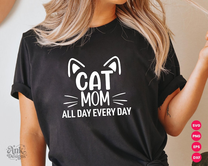 以下が含まれることがあります： 「CAT MOM ALL DAY EVERY DAY」の白文字グラフィックが入った黒いTシャツ。猫の耳とひげのデザインが含まれています。カジュアルなスタイルのTシャツです。