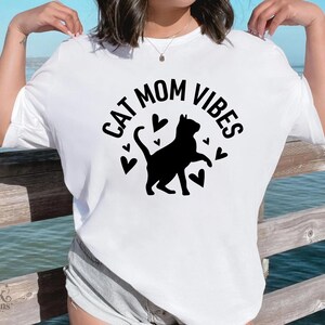 以下が含まれることがあります： 黒い文字で「CAT MOM VIBES」と書かれた白いTシャツ。黒い猫のシルエットとハートの形が特徴です。猫好きにぴったりのカジュアルなスタイルです。