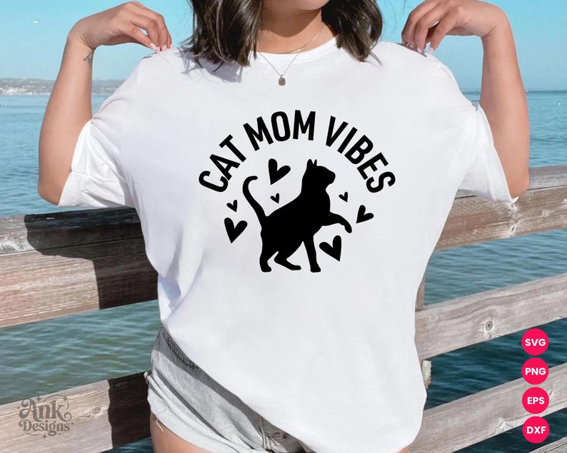 以下が含まれることがあります： 黒い文字で「CAT MOM VIBES」と書かれた白いTシャツ。黒い猫のシルエットとハートの形が特徴です。猫好きにぴったりのカジュアルなスタイルです。