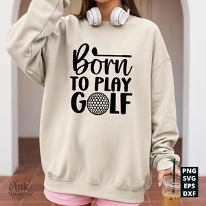 May include: A beige sweatshirt with the text "Born to Play Golf" in black. The design includes a golf ball and a golf club. The sweatshirt is a casual style, suitable for golf enthusiasts.