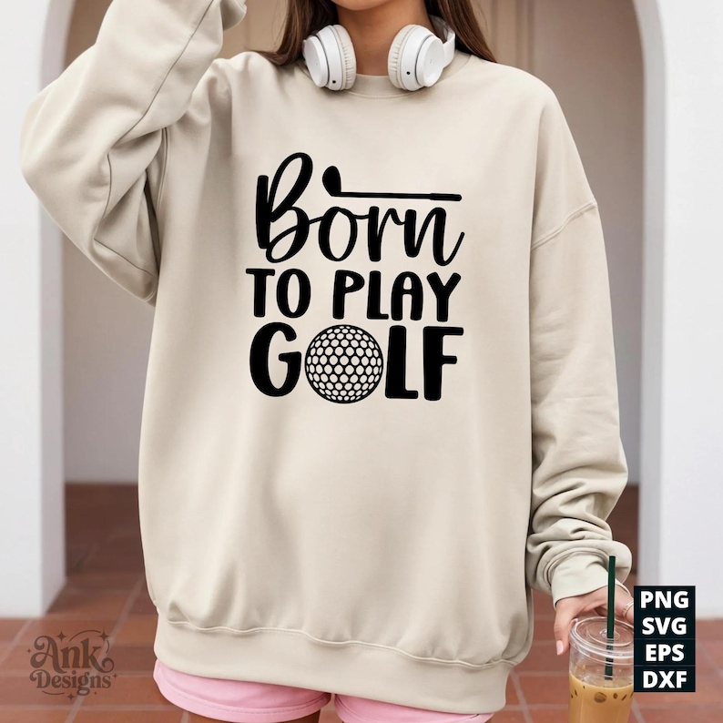 May include: A beige sweatshirt with the text "Born to Play Golf" in black. The design includes a golf ball and a golf club. The sweatshirt is a casual style, suitable for golf enthusiasts.