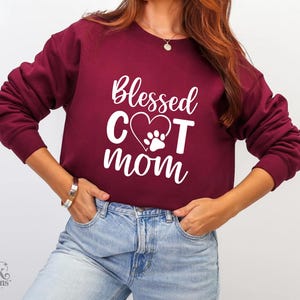 以下が含まれることがあります： 「Blessed Cat Mom」の白文字がプリントされた、マルーン色のスウェットシャツ。デザインには、足跡が入ったハートが含まれています。スウェットシャツはクルーネックです。